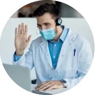 A healthcare professional in a lab coat and headphones waves while sitting at a laptop, engaged in a virtual consultation.