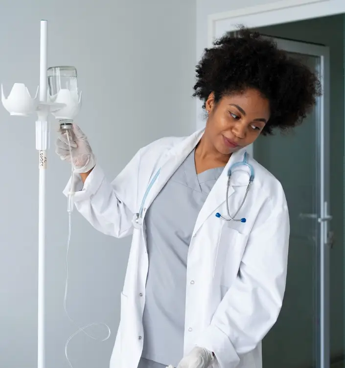 A healthcare professional in a white lab coat prepares an IV drip, wearing gloves and a stethoscope, in a modern medical setting.