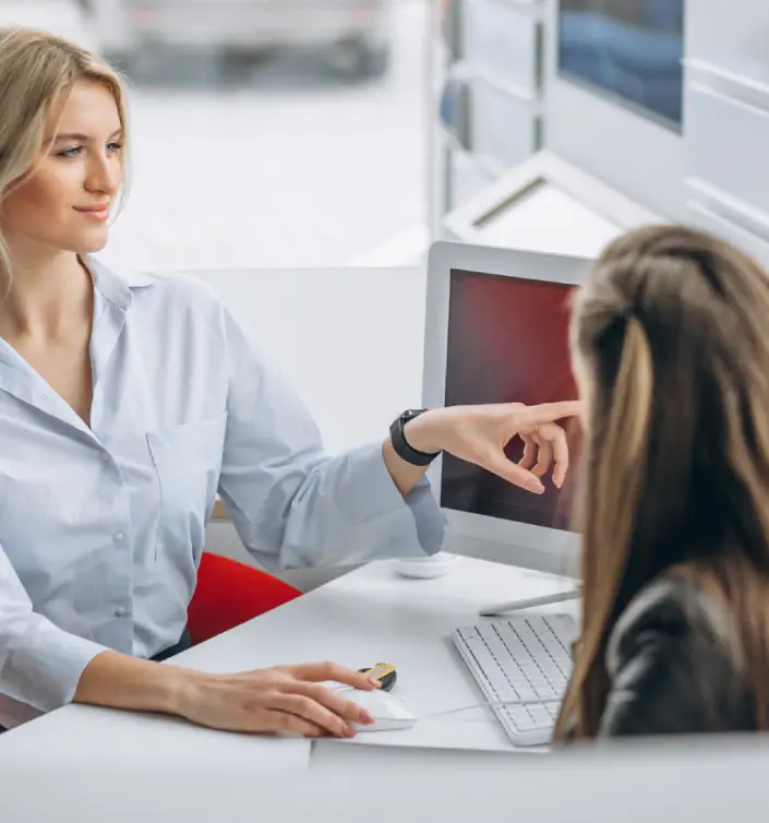 A woman in a light blue shirt gestures towards a computer screen while speaking to another seated individual.