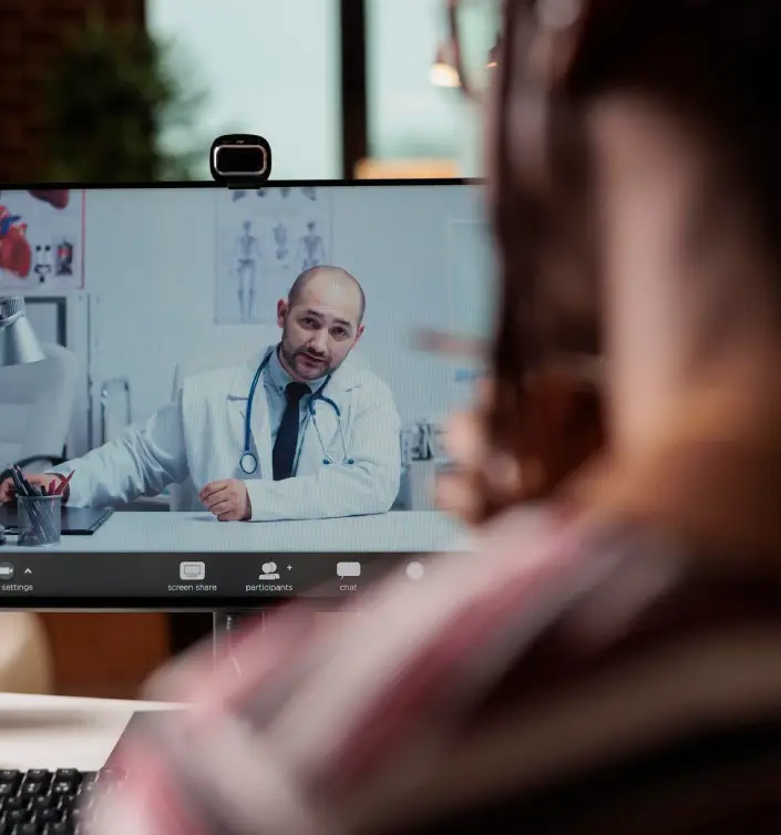 A person engages in a telehealth consultation with a doctor on a computer screen, featuring medical diagrams in the background.