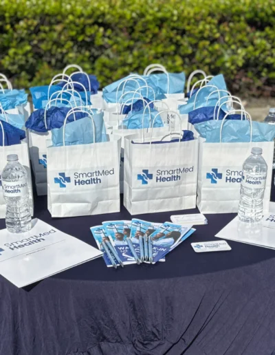 An outdoor promotional setup for SmartMed Health in a parking lot. A tall banner advertises "Walk-in Medical Care," next to a table with branded gift bags and water bottles.